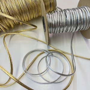 May include: Two spools of metallic trim, one gold and one silver, with the trim wound around the spools. The trim is a thin, flat cord with a subtle sheen.