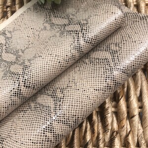 May include: A close-up of a beige snakeskin fabric with a subtle black pattern. The fabric is folded in half and is lying on a woven wicker surface.