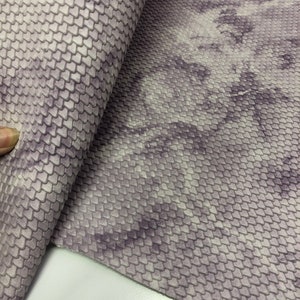 May include: A close-up of a purple and white faux leather fabric with a textured, scale-like pattern. The fabric is folded in half, revealing the pattern on both sides.