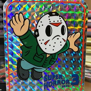 K&ouml;nnte beinhalten: Ein holografischer Aufkleber mit einer Comicfigur, die eine Hockeymaske und ein gr&uuml;nes Hemd tr&auml;gt. Der Aufkleber tr&auml;gt die Aufschrift "Super Horror 3" und "Pixel Bros."