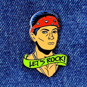 May include: A colorful enamel pin featuring a cartoon character wearing a red bandana with white stars and a green banner that says "Let's Rock!"