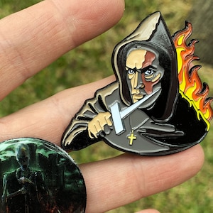 May include: Two enamel pins, one depicting a hooded figure holding a dagger with flames behind them, and the other depicting a dark figure with a sword in a swirling, fiery background.