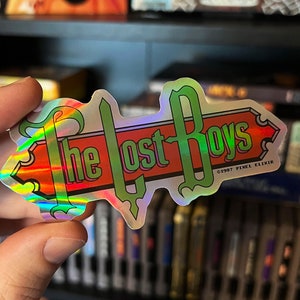 Könnte beinhalten: Ein holografischer Aufkleber mit dem Text "The Lost Boys" in einer grünen, roten und weißen Schriftart. Der Aufkleber hat einen silbernen Hintergrund und ein Copyright-Symbol mit dem Text "1997 Pixel Elixir".