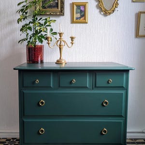 May include: A dark teal chest of drawers with gold handles and a candelabra. The chest has five drawers and is set against a white textured wall with framed artwork and mirrors. A potted plant sits on the chest.