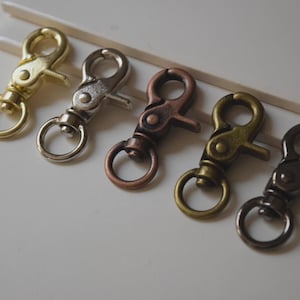 May include: Five metal swivel clasps in various finishes: gold, silver, copper, antique brass, and gunmetal. Each clasp has a ring and a lever for attaching to a strap or chain. The clasps are arranged diagonally on a white surface.