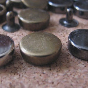 May include: Close-up of various metal snap fasteners in silver, bronze, and black. The fasteners are round with a flat top and a post on the underside. Some have a ring around the post. The fasteners are on a textured, brown surface.