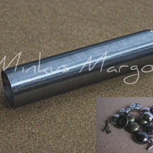 May include: A silver metal tube with a hollow centre, lying horizontally on a brown surface. The text "Minka Margo" is written in cursive. A collection of metal snaps and fasteners are in the lower right corner.