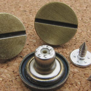 May include: Close-up of antique brass jeans buttons and rivets. Two round buttons with a horizontal groove, a rivet, and a screw are displayed. The hardware is on a corkboard surface.