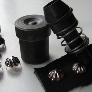 May include: A collection of silver-colored metal studs with a flower-like design, a black metal tool, and a black metal cylinder. The studs are scattered on a white surface, with two studs attached to a piece of black fabric.