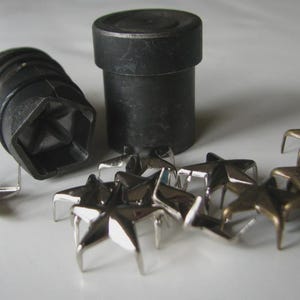 May include: A collection of metal star-shaped studs and two black metal tools. The studs have a silver finish and are arranged in a scattered pattern. The tools are cylindrical and have a dark, matte finish.