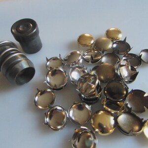 May include: An assortment of metal rivets and setting tools. The rivets are in various metallic finishes, including silver, gold, and dark bronze. The setting tools are cylindrical and made of dark metal.
