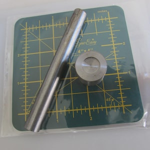 May include: A metal sewing tool set, including a cylindrical punch and a circular die, rests on a green cutting mat. The mat has grid lines and the text "Sew-Easy" printed on it. The tools are in a clear plastic bag.