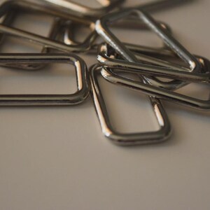 Op de afbeelding: Close-up van verschillende glanzende, zilverkleurige metalen rechthoekige en driehoekige ringen. De ringen liggen verspreid op een witte ondergrond, waarbij sommige elkaar overlappen. Het metaal heeft een reflecterende kwaliteit en vangt het licht.