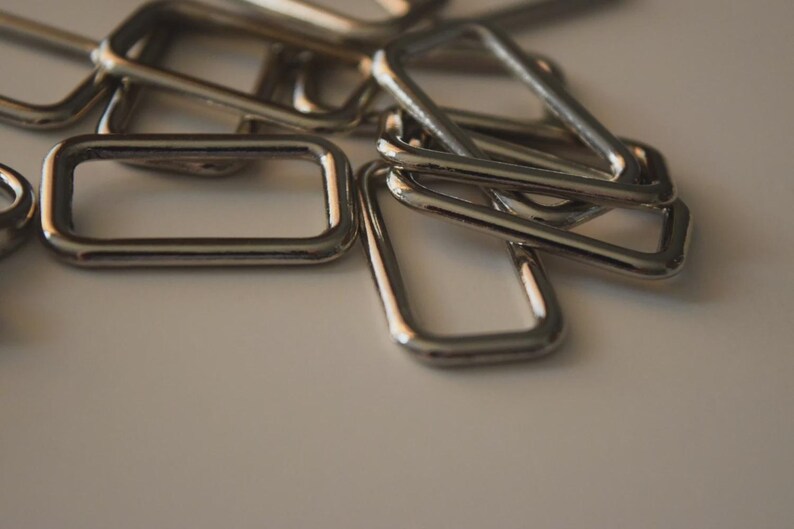 Op de afbeelding: Close-up van verschillende glanzende, zilverkleurige metalen rechthoekige en driehoekige ringen. De ringen liggen verspreid op een witte ondergrond, waarbij sommige elkaar overlappen. Het metaal heeft een reflecterende kwaliteit en vangt het licht.
