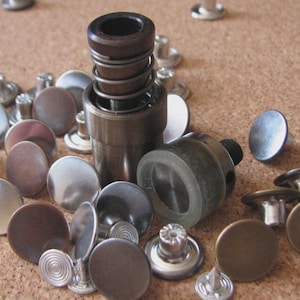 May include: A collection of metal jean buttons and a button setting tool. The buttons are in various colors, including silver, bronze, and brown. The setting tool is made of metal and has a spring mechanism.
