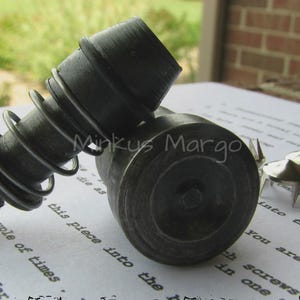 May include: Close-up of a dark gray metal object with a threaded end, a spring, and a cylindrical head. The object is lying on a piece of paper with text. Several small metal studs are also visible.
