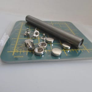 Peut inclure: Un ensemble de boutons-pression en métal et un outil de pose sont disposés sur un tapis de coupe vert. L'ensemble comprend divers composants de boutons-pression et un outil cylindrique, probablement pour fixer les boutons-pression.