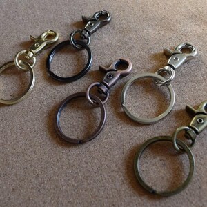 Puede incluir: Una variedad de llaveros con diferentes acabados metálicos, incluyendo dorado, negro, bronce, plateado y latón antiguo. Cada llavero cuenta con un cierre giratorio y un anillo circular. Los llaveros están dispuestos sobre una superficie de tono neutro.