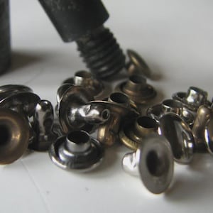 Peut inclure: Gros plan sur des rivets métalliques et un outil de pose. Les rivets sont de couleur argent et bronze, de formes et de tailles variées. L'outil de pose est gris foncé et a une extrémité filetée. Les articles sont éparpillés sur une surface blanche.