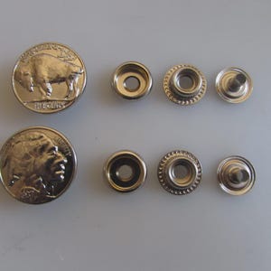 May include: A collection of silver-colored metal snaps and two buffalo nickel coins. The snaps include two sets of snap fasteners and four snap components. The coins feature a buffalo and a Native American profile.