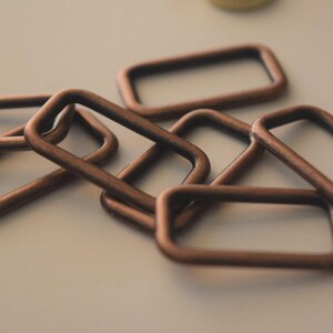 Op de afbeelding: Close-up van verschillende koperkleurige rechthoekige metalen ringen. De ringen zijn in een licht overlappend patroon gerangschikt, waarbij sommige plat liggen en andere schuin staan. Het metaal heeft een licht verouderd uiterlijk.