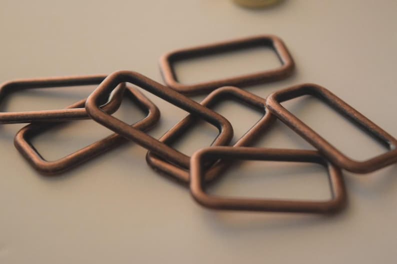 Op de afbeelding: Close-up van verschillende koperkleurige rechthoekige metalen ringen. De ringen zijn in een licht overlappend patroon gerangschikt, waarbij sommige plat liggen en andere schuin staan. Het metaal heeft een licht verouderd uiterlijk.