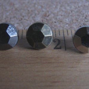 Puede incluir: Tres tachuelas metálicas facetadas en tonos bronce y plata se disponen sobre una superficie de madera junto a una regla. Las tachuelas tienen un diseño geométrico y probablemente se utilizan con fines decorativos.