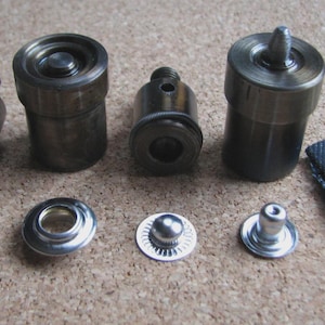 May include: An assortment of metal tools and hardware components, including cylindrical pieces, eyelets, and a snap fastener attached to a small piece of blue fabric. The items are in various sizes and shapes, with a metallic finish.