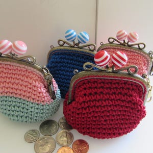 May include: Four crocheted coin purses with different colored yarn and metal clasps. Each purse has two striped buttons on top. The purses are arranged in a row with coins in front of them.