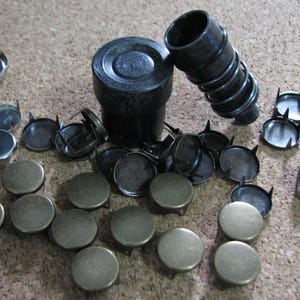 May include: An assortment of metal furniture tacks and a black tool for setting them. The tacks have a silver finish and a circular head. The tool is cylindrical with a textured grip. The items are scattered on a brown surface.