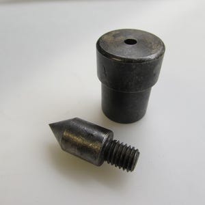 May include: Two metal tools, likely for leatherworking or similar crafts, are displayed against a white background. One is a cylindrical piece with a hole, and the other is a pointed, threaded tool. Both have a dark, aged appearance.