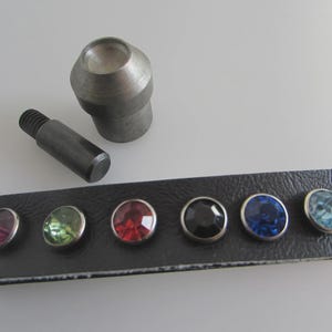 Könnte beinhalten: Eine Auswahl an farbigen Strassnieten in Rosa, Grün, Rot, Schwarz, Blau und Türkis wird auf einem schwarzen Lederstreifen gezeigt. Ein Metallwerkzeug und eine Schraube sind ebenfalls vorhanden.