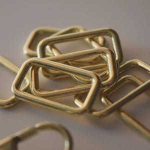 Puede incluir: Primer plano de varios anillos metálicos rectangulares de color dorado. Los anillos se superponen ligeramente, posiblemente para su uso en artesanía o como elementos decorativos. La iluminación resalta sus superficies lisas.