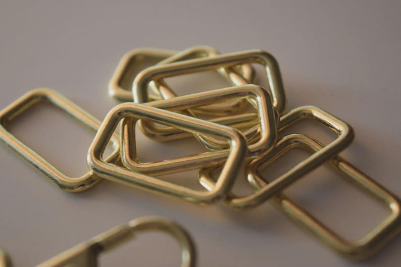 Op de afbeelding: Een close-up van verschillende goudkleurige, rechthoekige metalen ringen. De ringen overlappen elkaar licht, mogelijk voor gebruik in de ambacht of als decoratieve elementen. De belichting reflecteert op hun gladde oppervlakken.