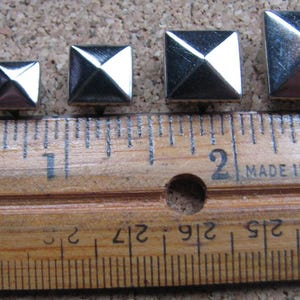 May include: Five silver-colored, square pyramid studs of varying sizes are arranged in a row. A wooden ruler with inch markings and the text "MADE IN U.S.A." is placed beneath the studs, providing scale. The studs have a shiny, reflective surface.