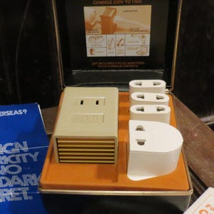 Może przedstawiać: Zestaw do konwersji energii elektrycznej z zagranicy w stylu vintage z wieloma adapterami wtyczek. Zestaw znajduje się w czarno-pomarańczowej walizce z niebieską broszurą z napisem "Foreign Electricity is No Deep Dark Secret."