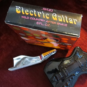 Może przedstawiać: Zestaw vintage Avon Electric Guitar Wild Country After Shave. Zestaw zawiera czarną butelkę w kształcie gitary, srebrną szyjkę gitary oraz pudełko z nazwą i wzorem produktu. Butelka zawiera 177 ml wody po goleniu.