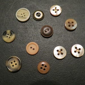 May include: A collection of various buttons in shades of brown, beige, and cream, scattered on a dark surface. The buttons vary in size, shape, and design, with two-hole and four-hole styles.