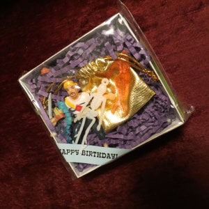 Puede incluir: Una caja de regalo con relleno de papel morado contiene una pequeña figura de pirata, una bolsa dorada y una figura de esqueleto translúcida. Se ve una pancarta azul con las palabras "HAPPY BIRTHDAY!".