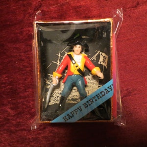 Puede incluir: Una figura de pirata en una caja decorativa. El pirata lleva una chaqueta roja, un sombrero negro y sostiene una espada. El fondo presenta un barco y una calavera. Una cinta azul dice "HAPPY BIRTHDAY!"