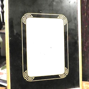 May include: Rectangular black picture frame with a gold border and a gold geometric design around the inner opening. The frame is made of a shiny material and is sitting on a wooden surface.