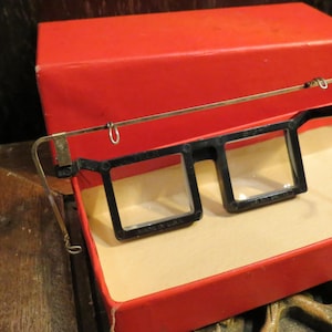 May include: Vintage black framed eyeglasses with clear lenses, resting inside a red box. The glasses have a metal bridge and arms. The box is open, revealing a beige interior. The glasses are likely from the mid-20th century.