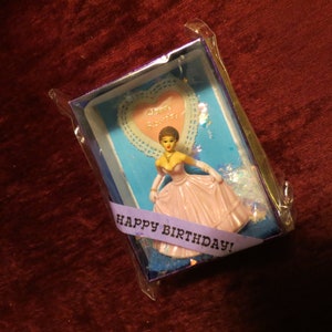 Puede incluir: Una caja de regalo de cumpleaños vintage con una figura de una mujer con un vestido rosa. La caja tiene un fondo azul, un encaje en forma de corazón y una cinta morada con "Happy Birthday!" escrito.
