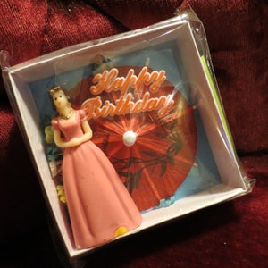 Puede incluir: Decoración de tarta de cumpleaños en caja. Presenta una figura con un vestido rosa, una sombrilla roja y la frase "Happy Birthday". La figura tiene un collar dorado y una tiara.