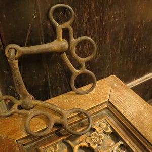 Puede incluir: Un antiguo bocado de caballo de metal desgastado, con un diseño complejo de múltiples anillos y articulaciones. El bocado descansa sobre una superficie de madera con detalles florales tallados, lo que sugiere un artículo ecuestre vintage.