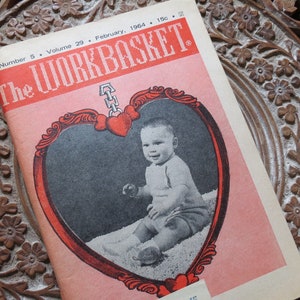 May include: A vintage copy of "The Workbasket" magazine from February 1964, with a baby on the cover inside a heart-shaped frame. The magazine rests on a carved wooden surface with floral patterns. The title is in red.