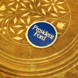May include: A round button with a blue background and the words "President Ford" in white. The button is resting on a wooden surface with carved floral designs.