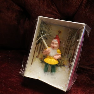 Puede incluir: Figura de gnomo de jardín en una caja. El gnomo lleva un gorro rojo, una camisa amarilla y pantalones verdes, sosteniendo un pico. La caja tiene un fondo de escena de bosque y está envuelta en plástico transparente.