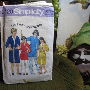 Può includere: Una busta di cartamodello Simplicity vintage con illustrazioni di persone in accappatoi. La busta è etichettata "8120 Simplicity" e "THE EVERY-BODY ROBES". L'illustrazione mostra quattro persone che indossano accappatoi blu, rossi e gialli.