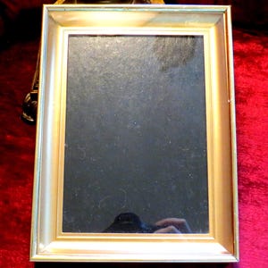 May include: A rectangular gold-coloured picture frame with a dark interior. The frame has a simple, elegant design with a slightly textured surface. The frame is set against a red background.